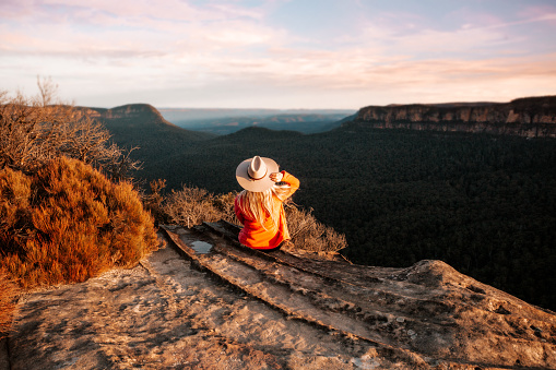 Woman sits on the edge of a cliff and looks out over the mountains in teh late afternoon