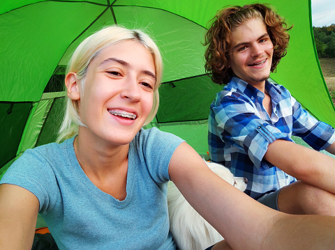 Happy young adult couple of campers taking selfie. Man and woman in casual clothes  with their cute white dog looking at camera. Love, leisure, technology concept
