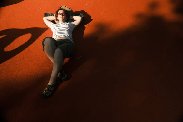 Active mid adult woman is relaxing on sunny sportsground holding her head on the skateboard
