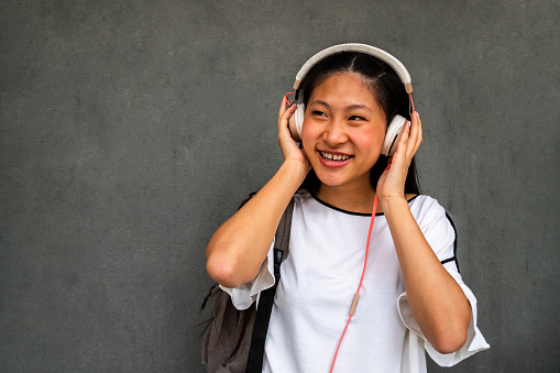 Smiling teen asian girl listening to music with headphones on dark concrete background. Copy space. Lifestyle concept.