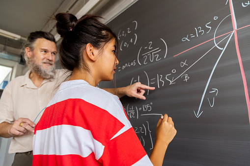 Mature caucasian male high school maths teacher explains blackboard exercise to asian girl student. Education concept.