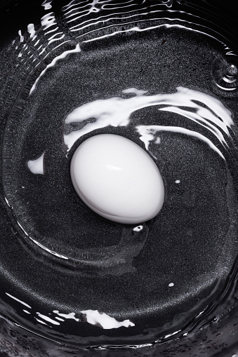 White egg in a water with circle waves on a black cooking pot background. Top view.