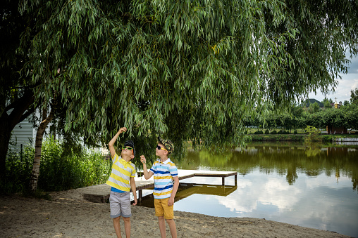 Siblings, brother, hugs. Two children, older boy and his younger brother standing on the shore of the lake near old tree. Summertime holiday concept leisure time on nature.