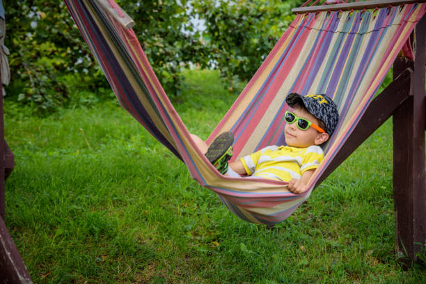 Cute little Caucasian boy relaxing and having fun in multicolored hammock in backyard or outdoor playground. Summer active leisure for kids. Child swinging on hammock. Activities for children.