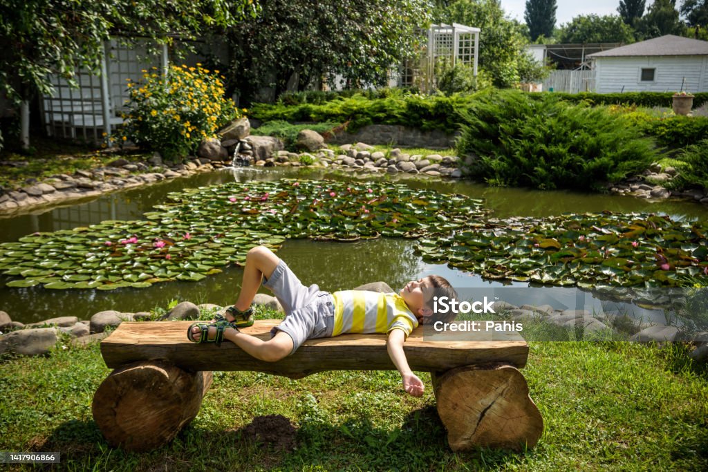 Boy is laying on a bench in a park near peaceful lake. Kid relaxing on nature on hot summer day. Clear blue sky Boy is laying on a bench in a park near peaceful lake. Kid relaxing on nature on hot summer day. Clear blue sky. 6-7 Years Stock Photo