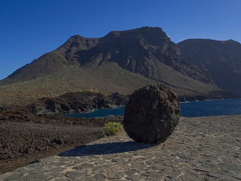 big rounded lava rock on paved path with view on hight lava cliffs los gigantes on Punta de Teno cape on west Tenerife canary island with blue sea and clear summer sky
