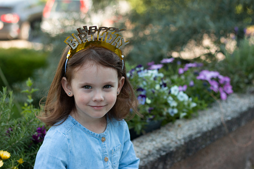 A little beautiful preschooler girl with a crown happy birthday on her head stands in a denim blue dress against the background of flowers