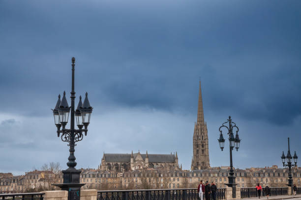 Selective blur on people walking in front of a bordeaux panorama and the tower of the Basilique Saint Michel basilica a cloudy afternoon. Picture of a cloudy panorama of Bordeaux, France, with the stone bridge, or pont de pierre, in front of the basilique saint michel steeple. fleche stock pictures, royalty-free photos & images