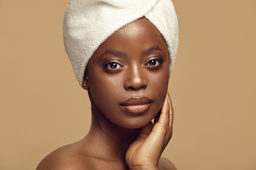Close up portrait of african american beautiful woman in spa head towel having healthy clean skin touching herself face on a beige isolated background.