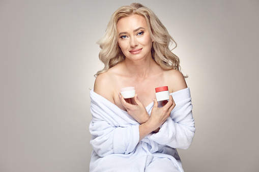 Good looking adult blonde woman with a clean and healthy skin wearing in spa bathrobe, holds variety regeneration creams with hydration for the face skin posing on beige isolated.