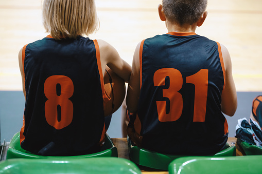 Two boys in basketball team sitting on substitute players bench. Children play basketball game. Youth basketball background. Sports education
