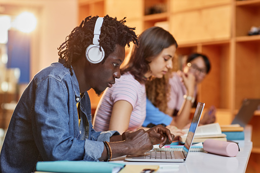 Diverse group of students in row using laptops and studying in college library, focus on black young man wearing headphones in foreground