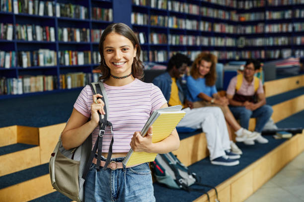 Portrait of Teenage Girl in School Library Waist up portrait of young woman with backpack standing in library and looking at camera, copy space boy and girl looking at camera stock pictures, royalty-free photos & images