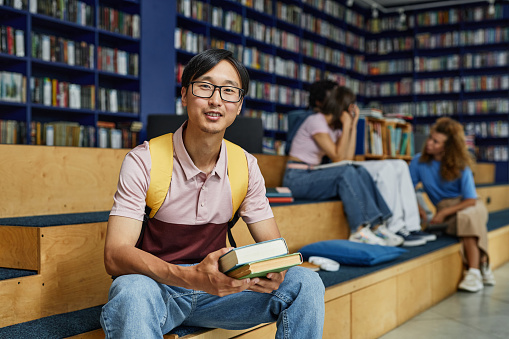A happy female student leaning on bookshelves at the library and smiling at the camera.