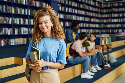 Waist up portrait of curly hair young woman holding books in college library and smiling at camera, copy space