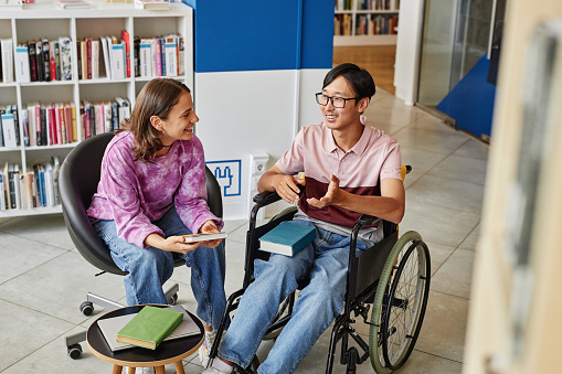 High angle portrait of Asian young man in wheelchair talking to friend while studying together in library