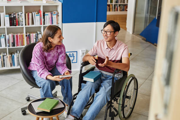 Young Student with Disability in College Library High angle portrait of Asian young man in wheelchair talking to friend while studying together in library 21 24 months stock pictures, royalty-free photos & images
