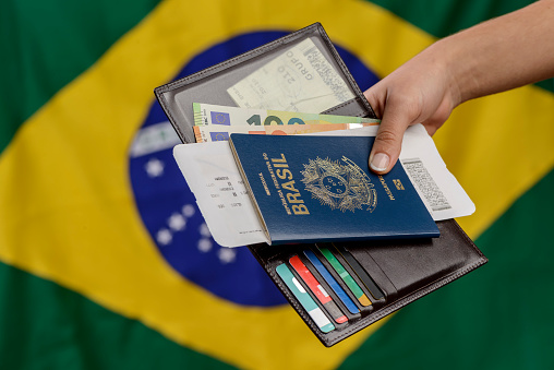 Hand holding Brazilian passport with Brazilian flag in the blurred background.