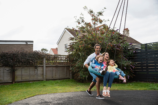 Full length side view of a father pushing his partner and his children in an outdoor play swing in a park in Seahouses in the North East of England. They all have beaming smiles on their faces as they have fun.