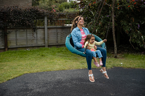 A full length side view of a woman who is playing in a playground with her young daughter. They are on a swing and she is holding on to her safely and encouraging her hold on to the chains in the park in Seahouses in the North East of England.