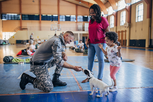 Diverse group of people, soldiers on humanitarian aid to civilians in school gymnasium, after natural disaster happened in city. Soldier with mother and daughter.