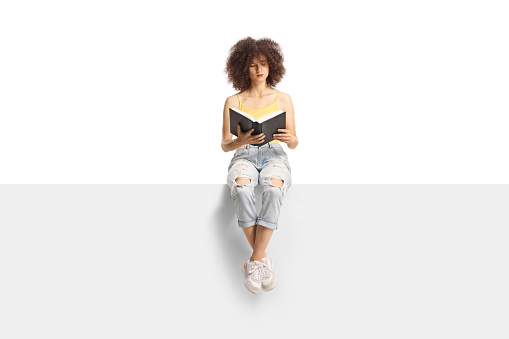 Young woman with curly hair sitting on a panel and reading a book isolated on white background