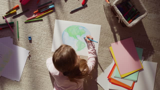 Top View: Little Girl Drawing Our Beautiful Planet Earth. Very Talented Child Having Fun at Home, Imagining Our Home Planet as a Happy Place with Clean, Sustainable Living. Cozy Sunny Day. Zoom out
