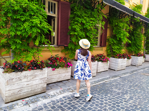 Rear view of young woman walking on the street of Lviv. Beautiful girl in white blue dress and straw hatenjoying visiting Lviv. Summer vacations.