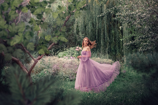 A girl in a long lilac dress stands in a clearing among the grass. In the background, trees, forest. The girl holds a bouquet of eustoma in her hand. Summer, nature, greenery.