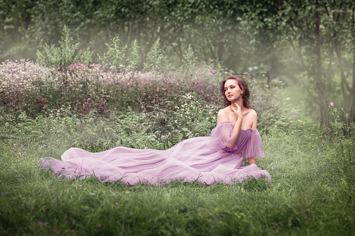 The girl is sitting in a clearing among the grass, standing in a long lilac dress. In the background are trees, forest and flowers. The girl raised her hand to her face. Summer, nature, greenery.