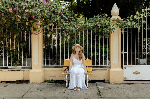 Girl sitting on a park bench. This bench is on the sidewalk in front of a house. The girl is wearing a white dress and a hat.