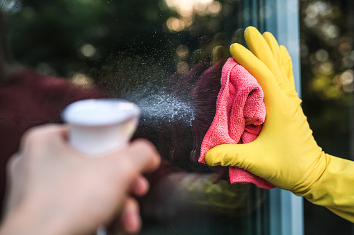 A woman's hands in yellow protective gloves cleaning a window pane with a pink rag and  a detergent in a spray bottle. Copy space.  Home routine and housekeeping