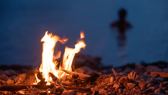 Alone man bathing in the river near the small campfire with gentle flames beside a lake during blur hour.