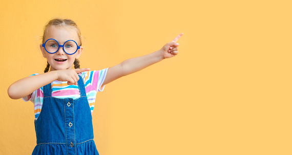 Special Offer. Smiling adorable girl in eyeglass pointing at copy space over yellow background. Colored striped clothes