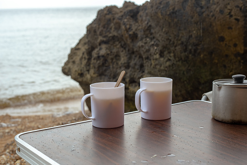 There are mugs of hot tea and a ladle on the tourist table. Sea coast with rocky shore on a rainy day, a place for recreation and tourism.