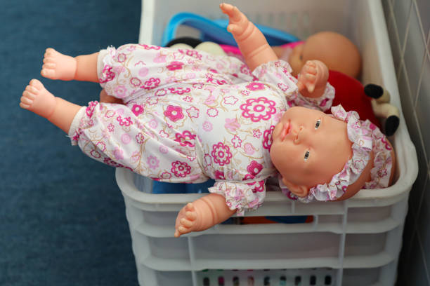 Back view of young woman holding realistic cute doll of newborn caucasian baby looking at camera. Unrecognizable girl sitting at table with reborns indoors. Selective focus on toy's face.