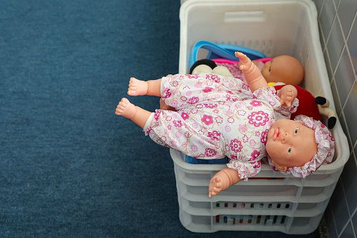 White plastic box on an oil blue floor with several plastic dolls inside. Coming out one of them with clothes in white and pink.