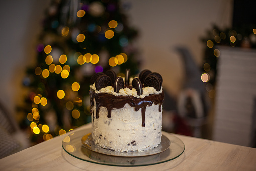 White cake with black cookies and with defocused christmas tree background. Selective focus. Food for celebrating New Year at cosy home.