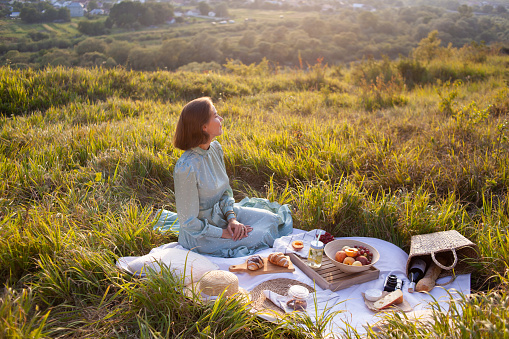 A girl in a long summer dress with short hair sitting on a white blanket with fruits and pastries and enjoying the view. Concept of having picnic in a city park during summer holidays or weekends.