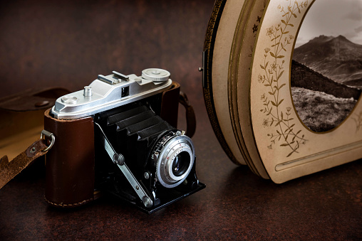 Antique folding bellows camera with leather strap and leather antique album. There is another small bellow camera in the background. The bellow camera in the foreground is from the 1930s and the bellow camera in the background is from 1960s.