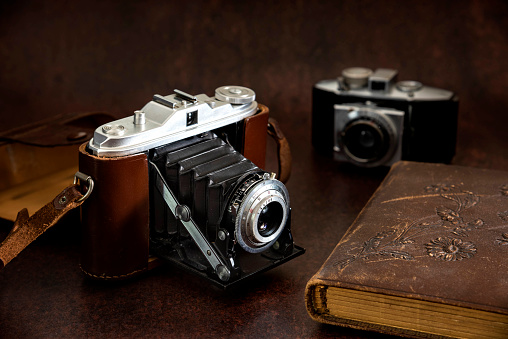 Antique folding bellows camera with leather strap and leather antique album. There is another small bellow camera in the background. The bellow camera in the foreground is from the 1930s and the bellow camera in the background is from 1960s.