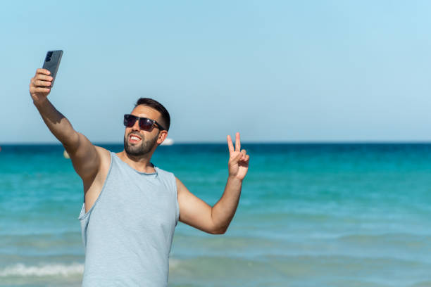 istock Young man with beard and sunglasses taking a selfie with his smartphone on the beach on a sunny day with plenty of copy space. 1417973873