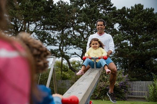 An over the shoulder view of a family of four playing on a seesaw in a public playground in Seahouses in the North East of England. The father is sitting with his daughter as they sit opposite his wife and daughter.