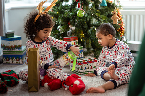 Two young siblings playing with their new gifts in front of a Christmas tree on Christmas morning. They are wearing matching pyjamas and are in the North East of England.