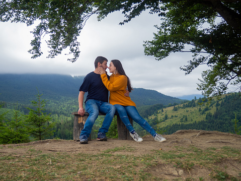 Young man and woman admiring breathtaking view while sitting on bench in the mountains. Lovely couple in casual clothes sit under large tree with forest background.