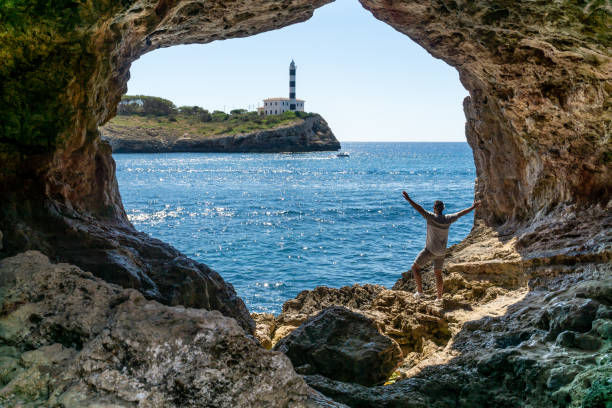 istock Unrecognizable man with raised hands inside a cave overlooking the sea and a red and white lighthouse with a ship sailing 1417978343