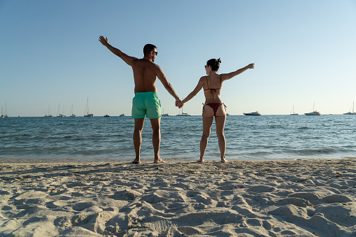 Couple on the shore of the beach with their hands raised and the boats in the background on a sunny day