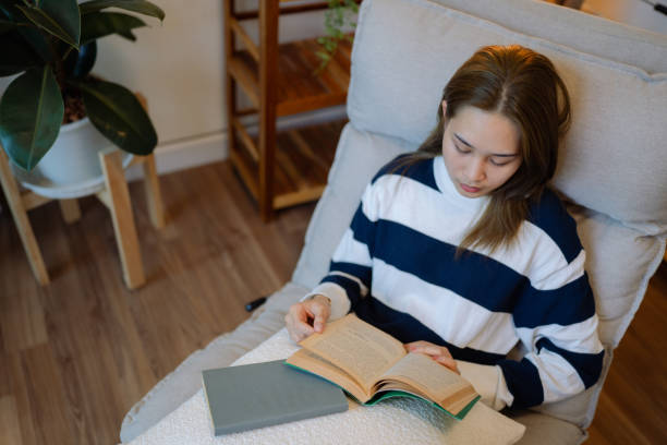 istock Asian woman reading a book on the sofa, she uses a tablet to write content. 1417980459