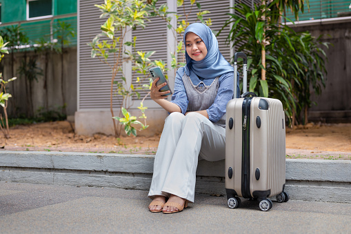 Young female Muslim traveler with hijab using smart phone to call for crowdsourced taxi while sitting with her wheeled luggage on pedestrian walkway.