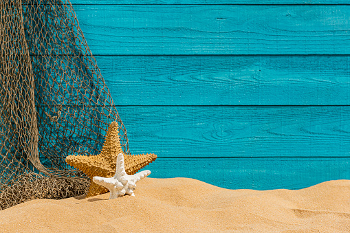 A turquoise colored raw sawn wooden plank wall, sand, fishing net and 2 starfish background